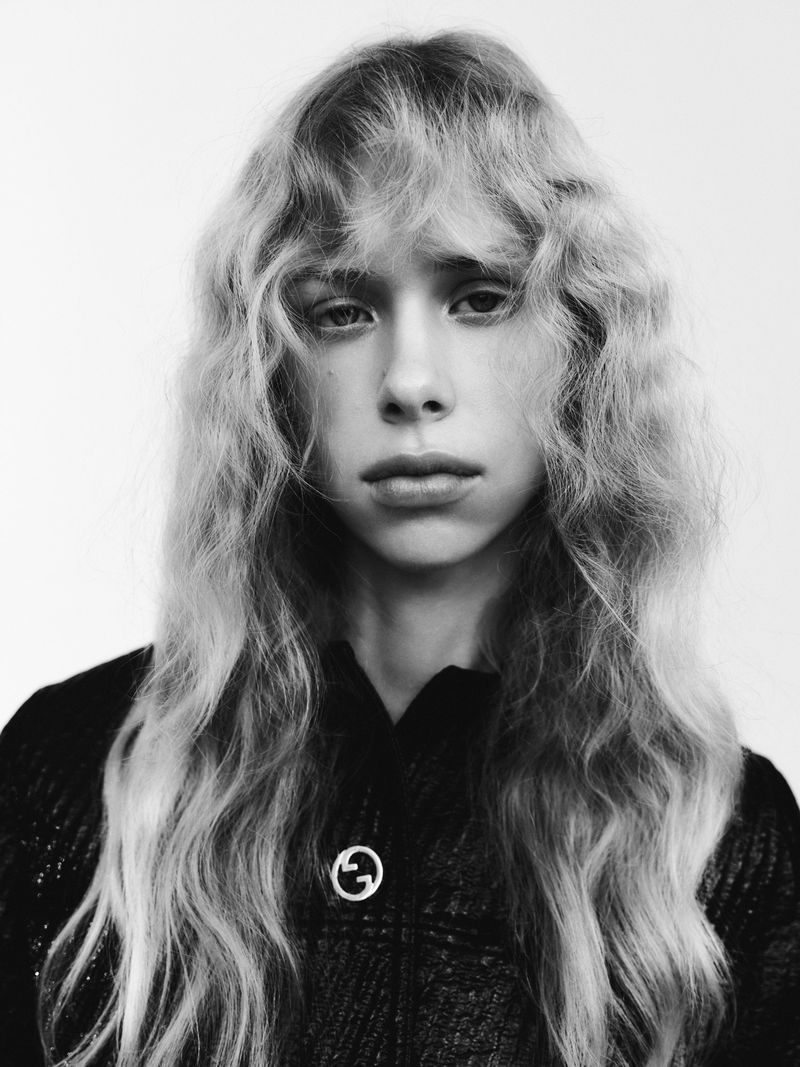 Monochrome portrait of a young fair-skinned person with long, wavy blonde hair and a black textured jacket indoors against a white backdrop.