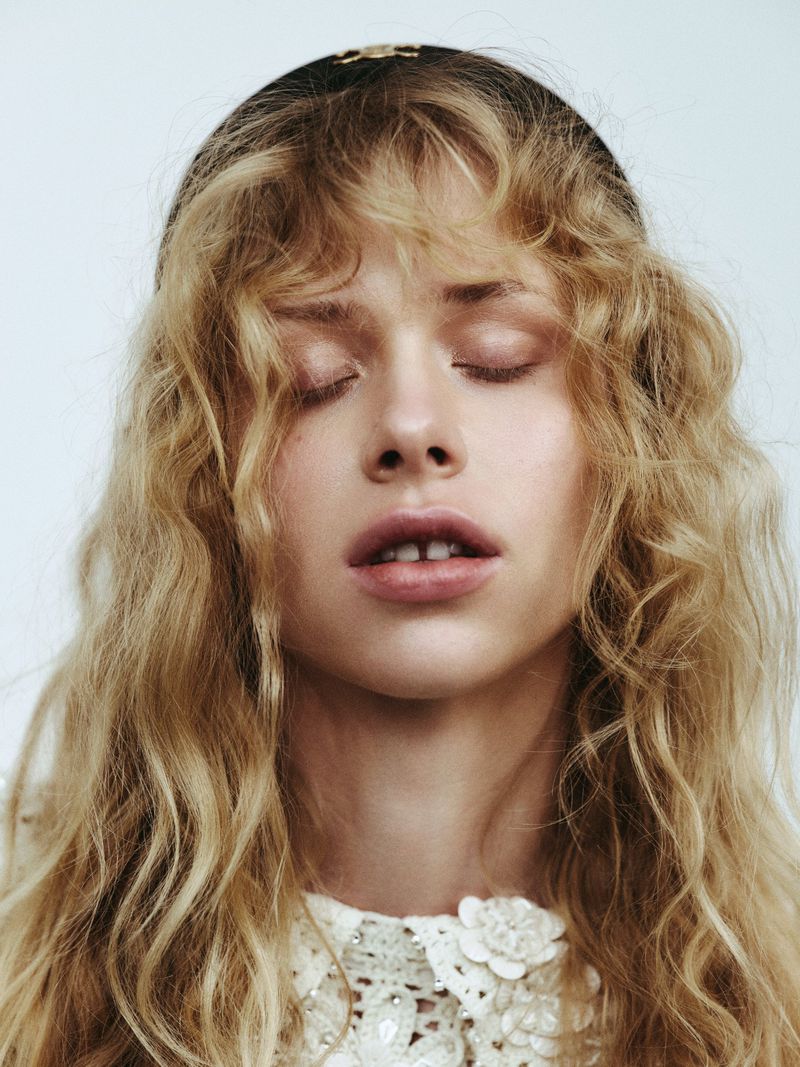 Close-up portrait of a young fair-skinned woman with wavy blonde hair and closed eyes, wearing a lacy white top and a dark headband.