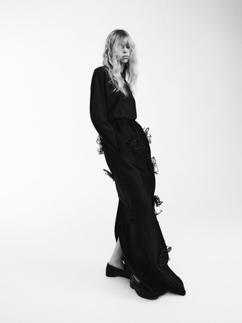 Black-and-white fashion shot of a single female model in a long sleeve top and maxi skirt with butterfly accents, standing in a bright studio.