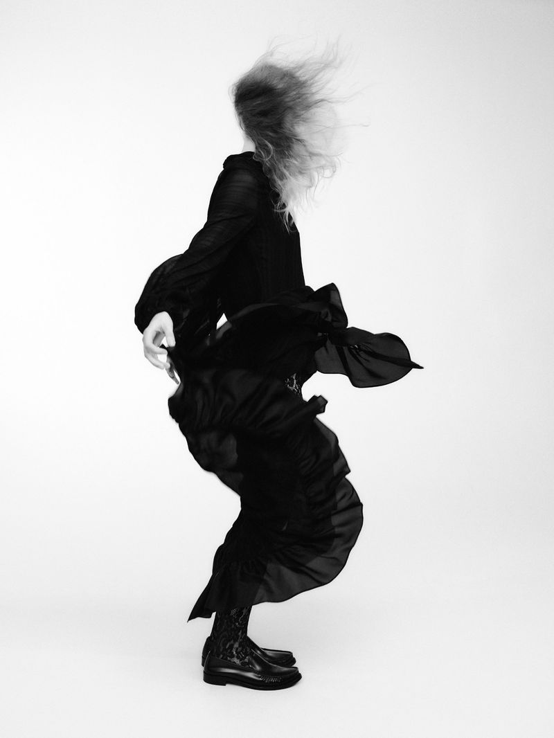 Monochrome fashion shot of a person in motion wearing a long, flowing dress and boots on a light background in a photo studio.