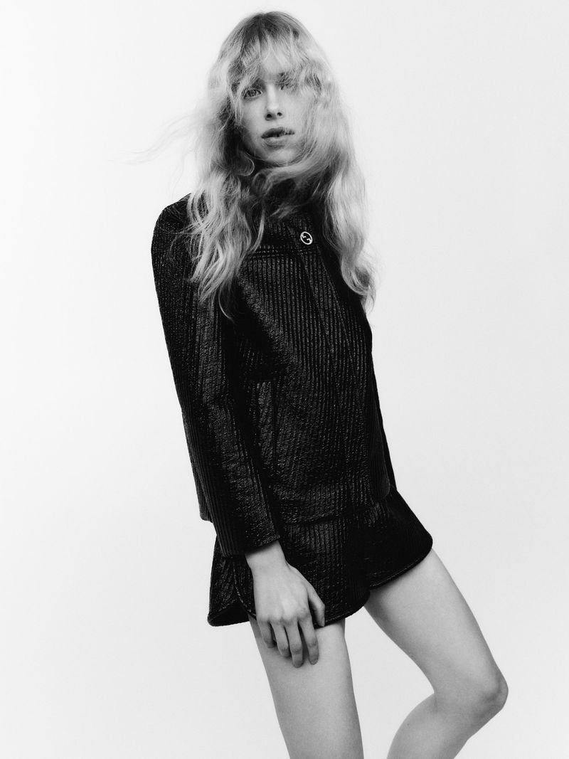 Model with long blonde hair wearing a textured dark jacket and shorts against a white background