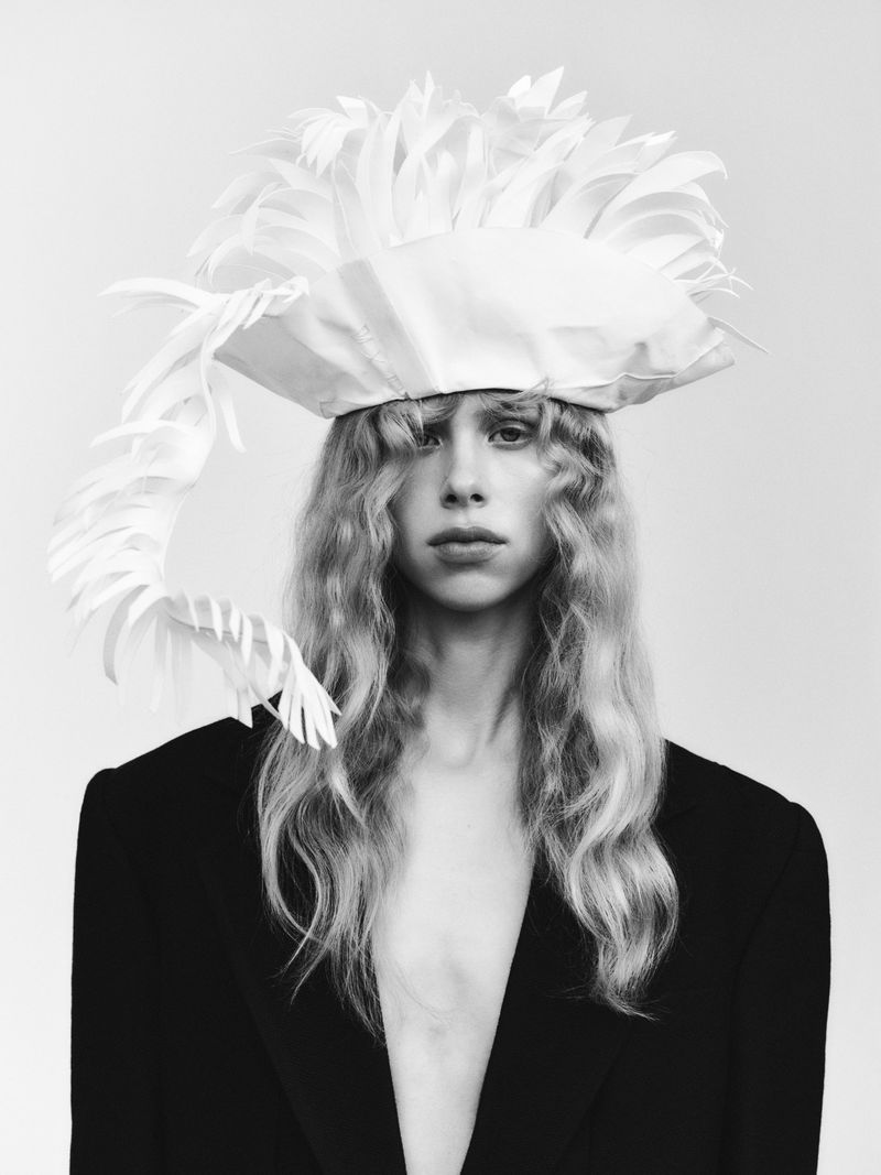 A person with long wavy hair wears a white feathered hat and oversized black jacket against a plain white background in a studio shot.