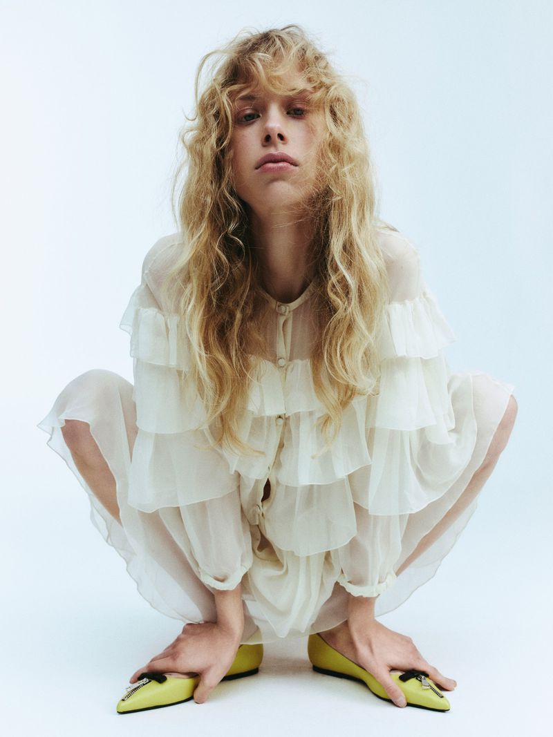 Fashion portrait of a fair-skinned person with curly blonde hair wearing a ruffled white dress and yellow flats against a white backdrop.