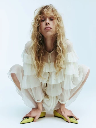 Fashion portrait of a fair-skinned person with curly blonde hair wearing a ruffled white dress and yellow flats against a white backdrop.