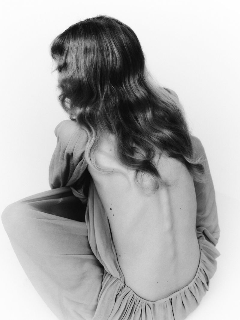 Monochrome photo of a slender, back-facing adult woman with long wavy hair in a floor-length dress, posing in a brightly lit photo studio.