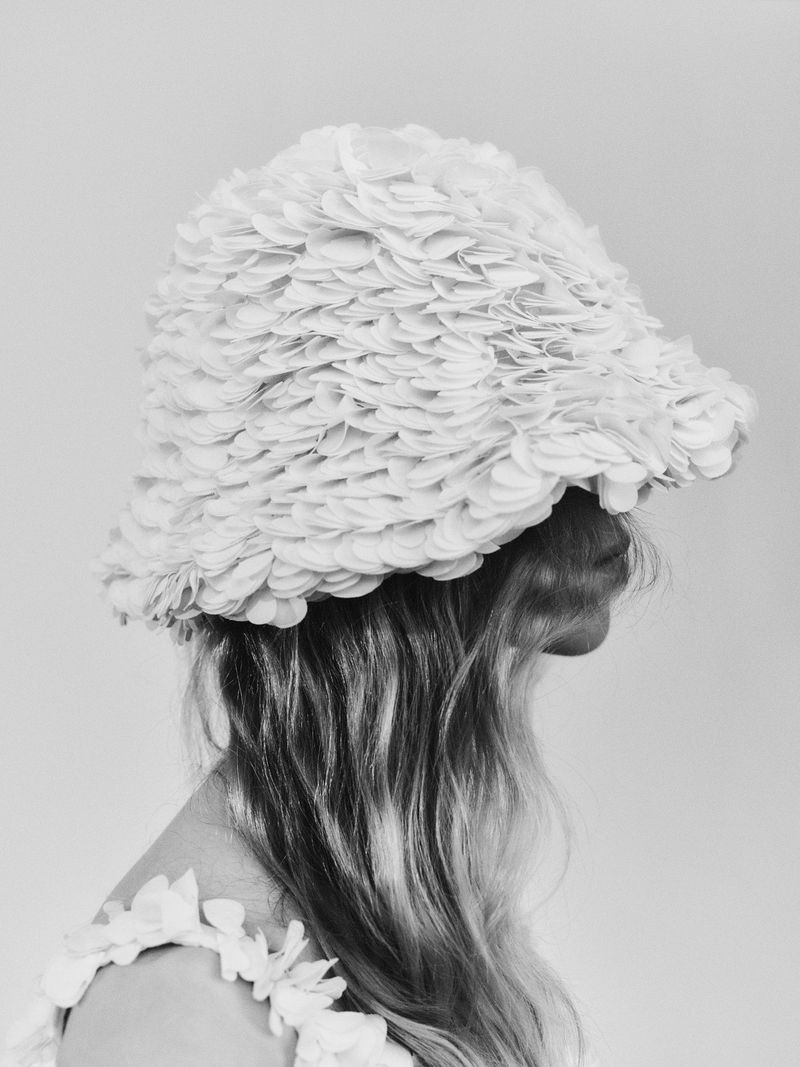 Black and white studio shot of woman in hat and top with flower appliques, looking away from camera, with light, neutral background.