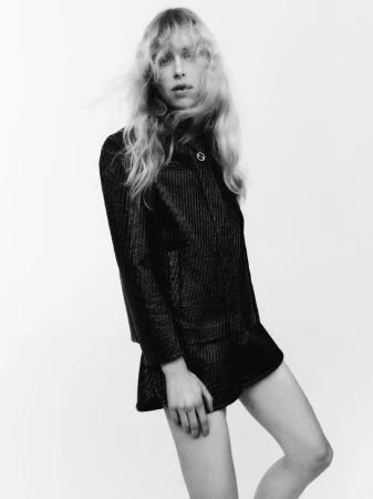 Model with long blonde hair wearing a textured dark jacket and shorts against a white background