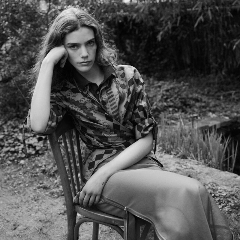 A young adult woman with long hair poses while sitting on a wooden chair outdoors wearing a patterned shirt and long skirt in a black and white photograph.