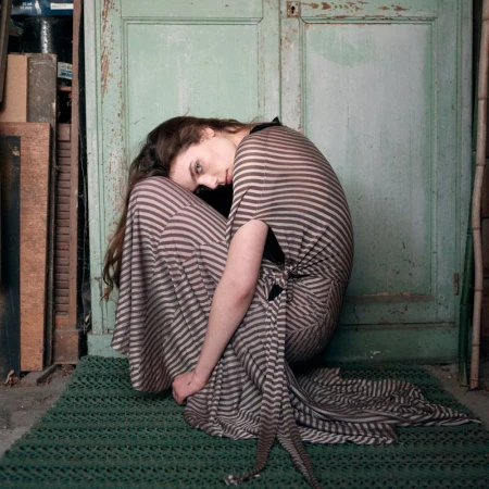 Brunette woman in striped dress sitting on green mat against weathered door. Portrait shot, indoor setting, soft lighting.