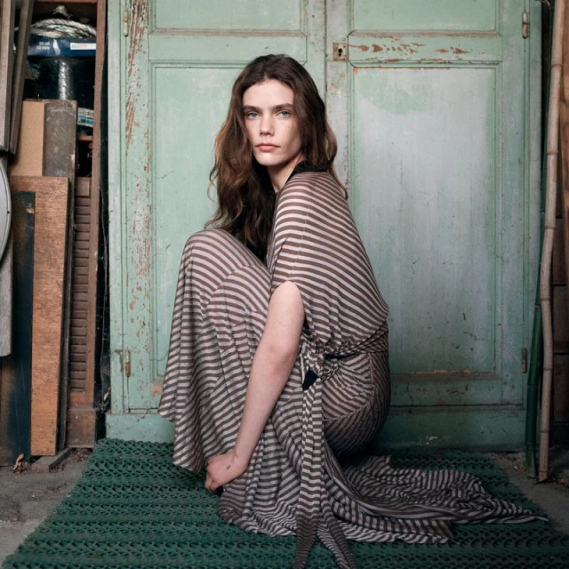 A woman with long, wavy brown hair poses in a striped maxi dress. She's sitting on a green woven mat in front of weathered green doors.