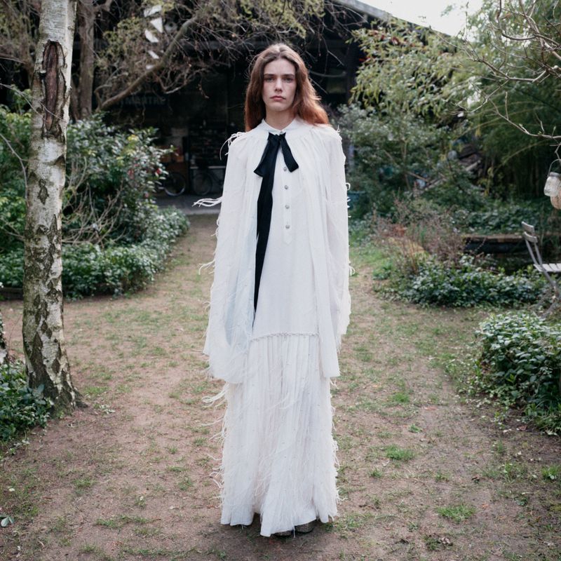Model standing on a grassy path wearing a white fringed dress with a black bow tie against a garden background.