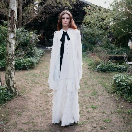 Model standing on a grassy path wearing a white fringed dress with a black bow tie against a garden background.