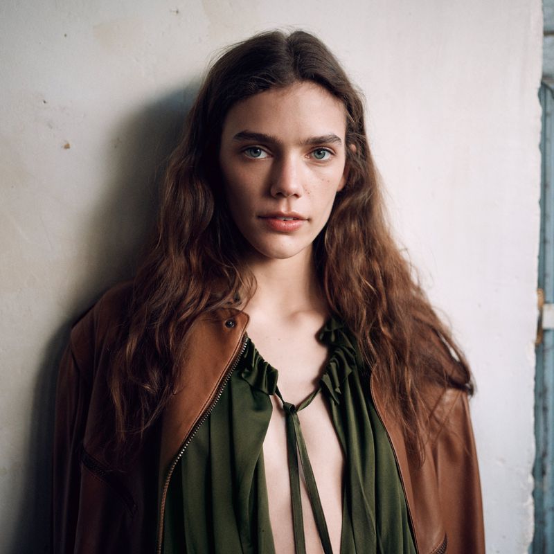 A young adult woman with wavy brown hair wears a brown leather jacket and green top in a close-up studio portrait against a white wall.
