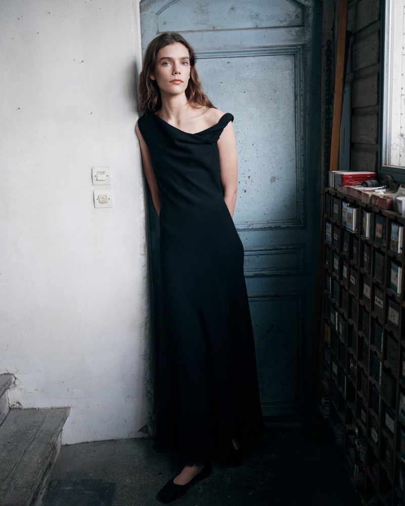 A woman in a black dress poses indoors, leaning against a blue door in a fashion shot, showcasing minimalist elegance and style.