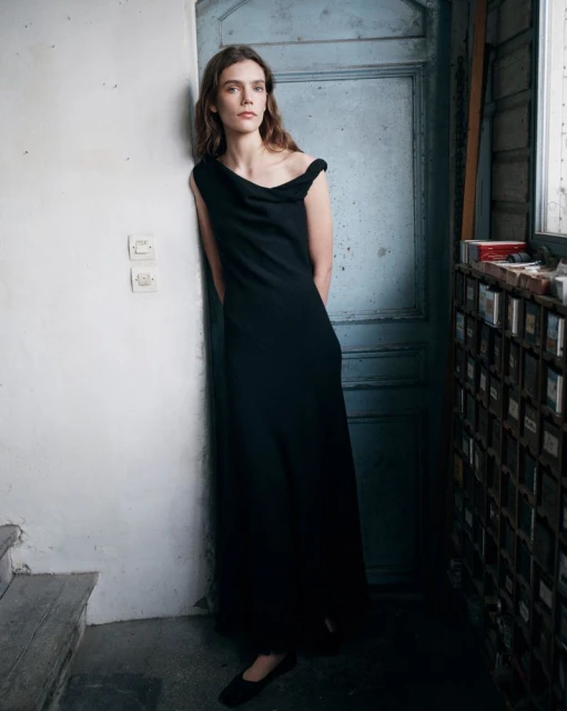 A woman in a black dress poses indoors, leaning against a blue door in a fashion shot, showcasing minimalist elegance and style.