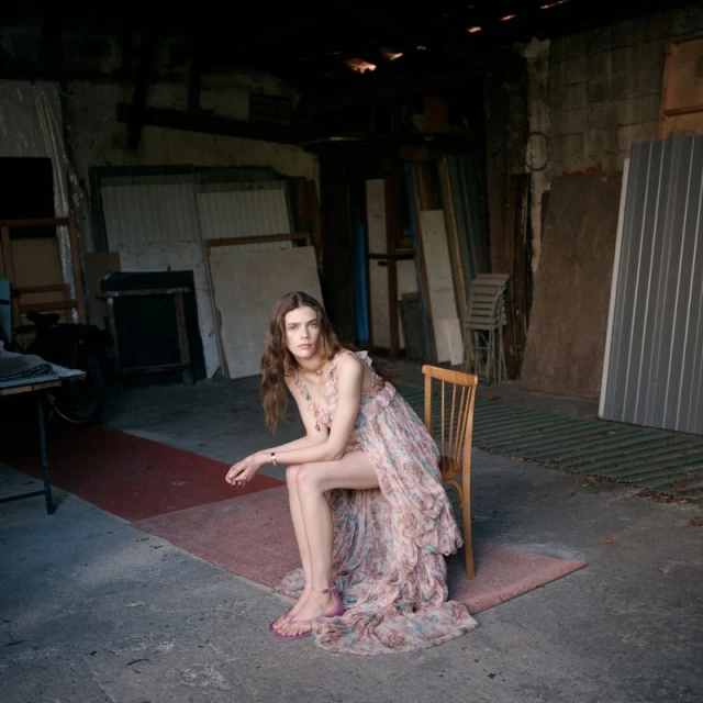 A young adult female model with long brown hair poses while seated on a wooden chair in a flowing floral dress in a dim indoor space.