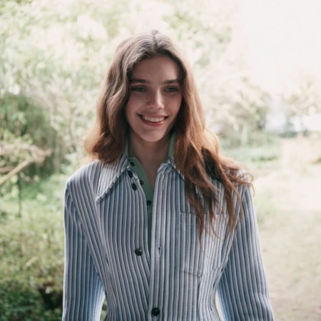 Portrait of a smiling woman with long brown hair wearing a striped blue and white shirt outdoors in a green natural setting.