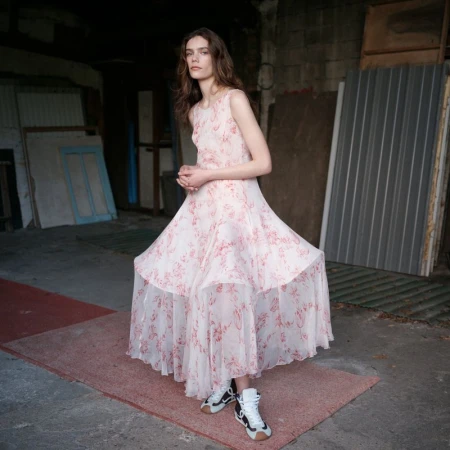 Woman in a flowing floral dress posing in an indoor space with sneakers, casting a contemplative gaze toward the viewer, fashion editorial.