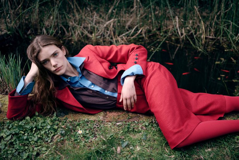 Woman in a red suit lies on a grassy patch near a pond with goldfish, showcasing an editorial fashion shot with natural lighting.