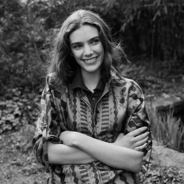 Black-and-white medium shot of a smiling woman with her arms crossed, wearing a patterned shirt, outdoors in a wooded environment.