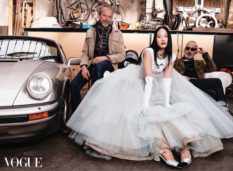 Editorial fashion shot of an Asian woman in a white dress sitting with two men and a classic Porsche in a garage-like setting. Vogue logo visible.