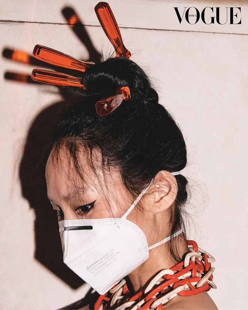 A person with dark hair in an updo with orange hair sticks, wearing a mask, and a red and white chain necklace, Vogue logo visible.