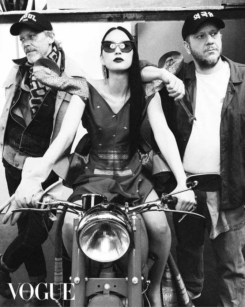 Editorial fashion shot of a model seated on a motorcycle with two adult males standing behind in an indoor studio setting in black and white.
