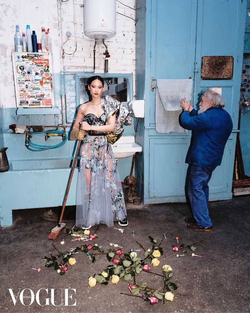 Editorial fashion shot of an Asian woman in a gown standing in a washroom with a bearded man adjusting a towel and flowers on the floor.