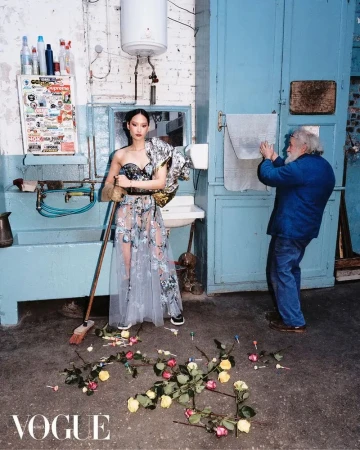Editorial fashion shot of an Asian woman in a gown standing in a washroom with a bearded man adjusting a towel and flowers on the floor.