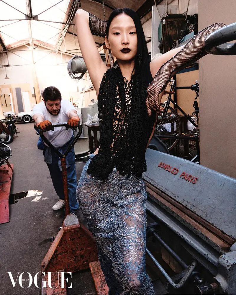 Asian woman poses in a black top and silver skirt in a garage while a man pushes a dolly with equipment in the background.