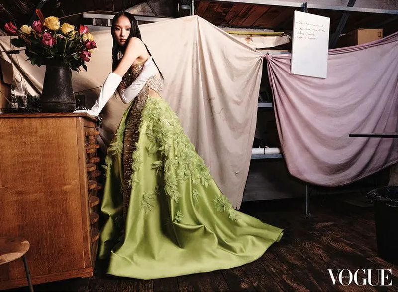 Woman poses in a green couture gown with flowers, standing in front of a draped backdrop and wooden cabinets in studio setting.