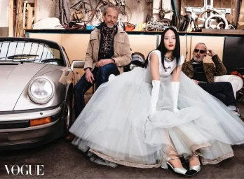 Model in voluminous white gown and opera gloves seated beside a silver car in a workshop filled with bicycle parts and two men