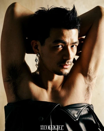 Portrait of a man with arms raised wearing a black leather jacket and dangling earring against a neutral wall.