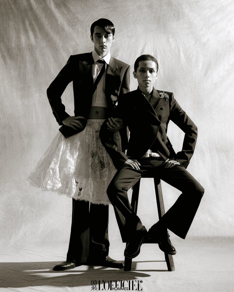 Two men pose in formal black suits and a white skirt in a studio setting, showcasing a fashion-forward and gender-fluid aesthetic.