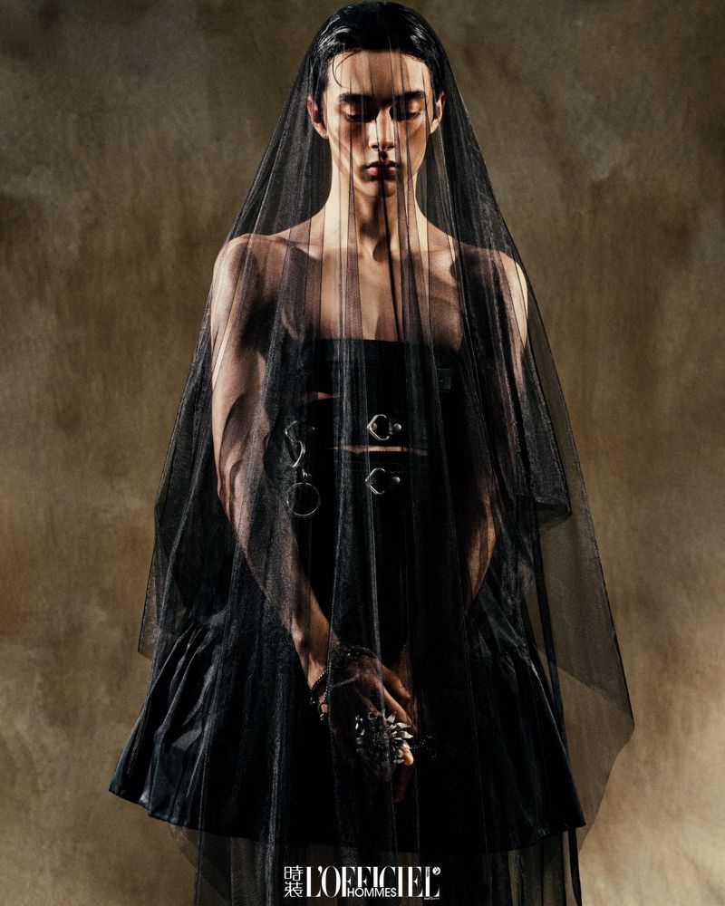 Fashion editorial portrait of a slender male model in a black dress and veil, posing in a photo studio against a textured background.