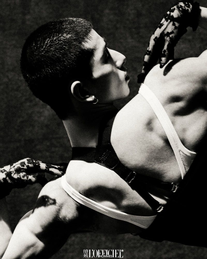Monochrome fashion shot features a man with a short haircut in a harness and underpants, with gloved hands touching him, on a dark background.