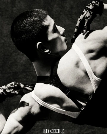 Monochrome fashion shot features a man with a short haircut in a harness and underpants, with gloved hands touching him, on a dark background.