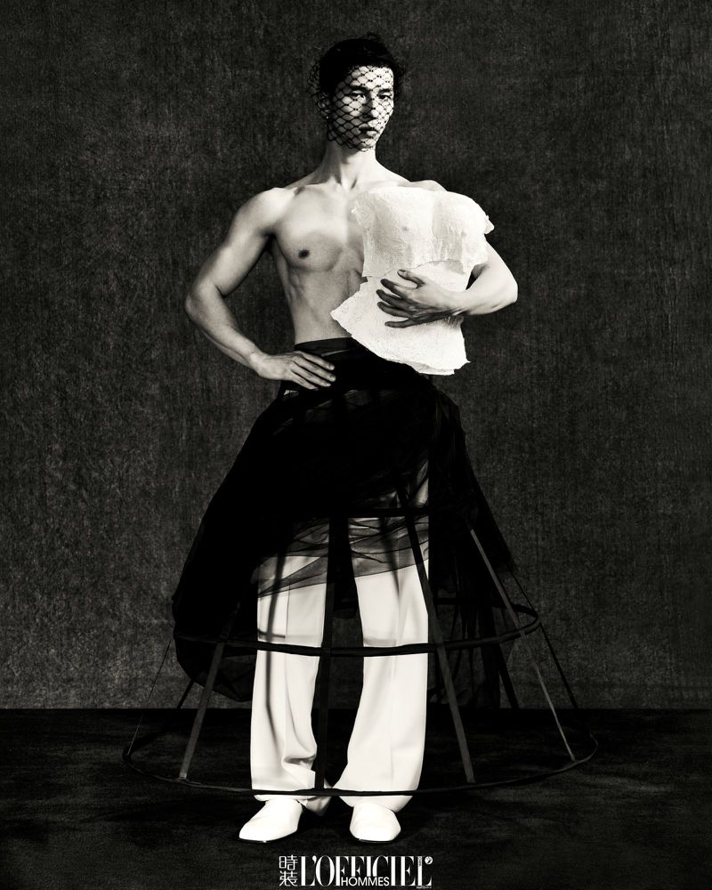 Male fashion model in avant-garde attire poses against a textured backdrop, featuring a sheer face veil, deconstructed top, and cage skirt.