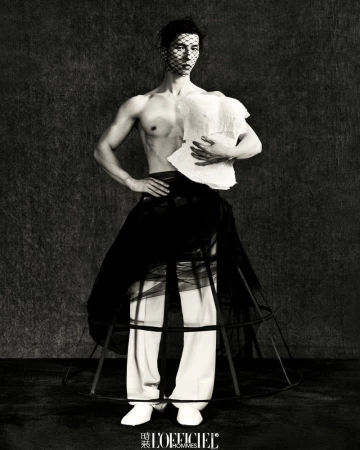 Male fashion model in avant-garde attire poses against a textured backdrop, featuring a sheer face veil, deconstructed top, and cage skirt.