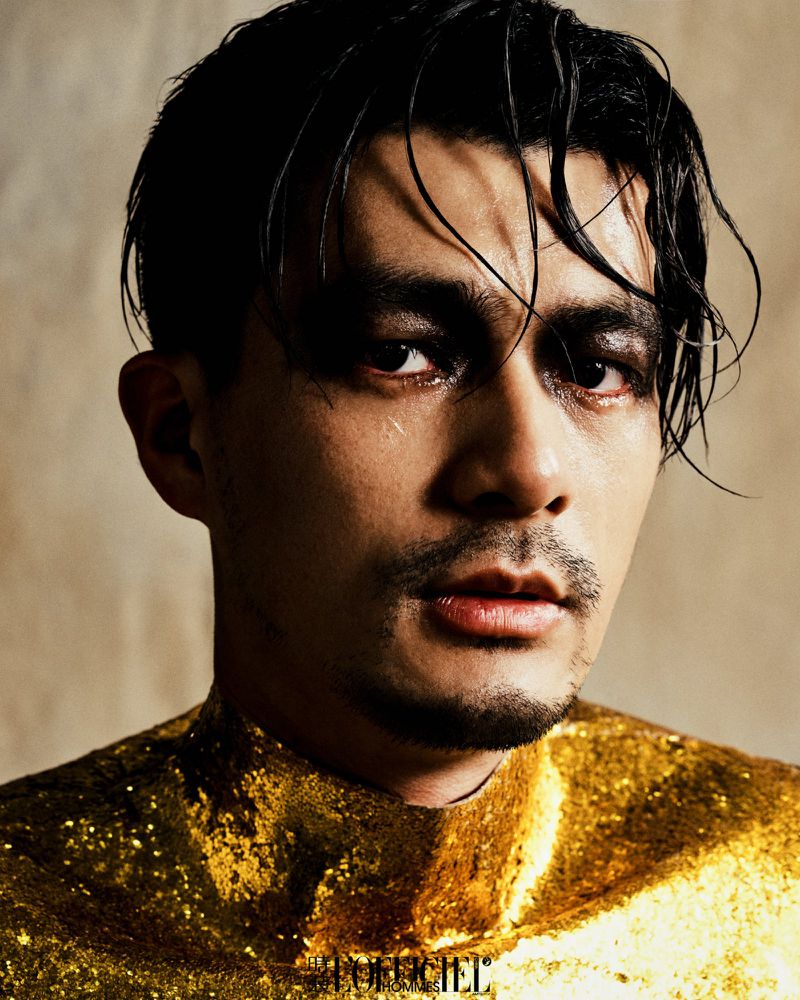 Close-up portrait of an adult male with dark hair and dramatic makeup, wearing a gold sequined top, against a neutral background.
