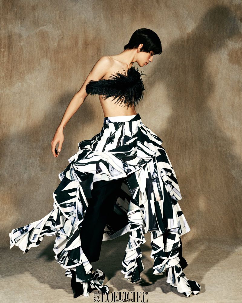Fashion editorial shot of a person in a black feathery top and a large, ruffled black-and-white geometric skirt against a brown backdrop.