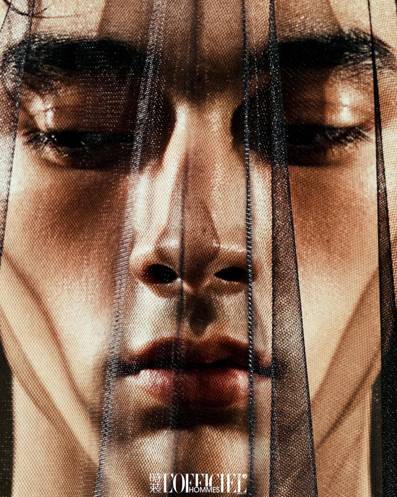 Close-up portrait of a young adult male with light skin, partially veiled with a thin black net, in soft light with a magazine logo.