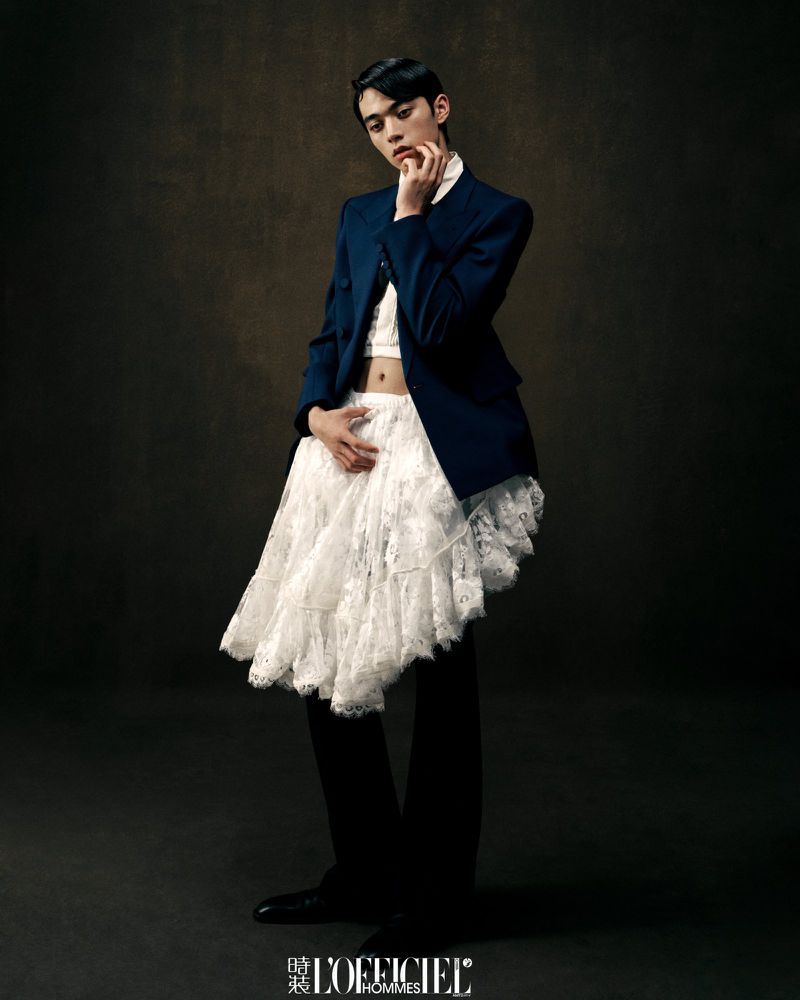 A person with dark hair and wearing a navy blazer, white top and white lace skirt poses against a dark background in a studio shot.