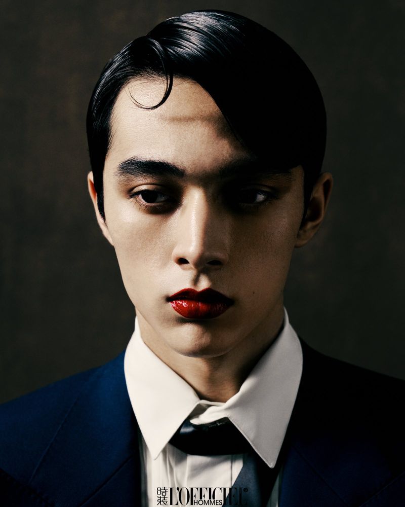 Dramatic portrait of a young male with dark hair wearing red lipstick, a white dress shirt, dark tie, and suit jacket against a grey background.