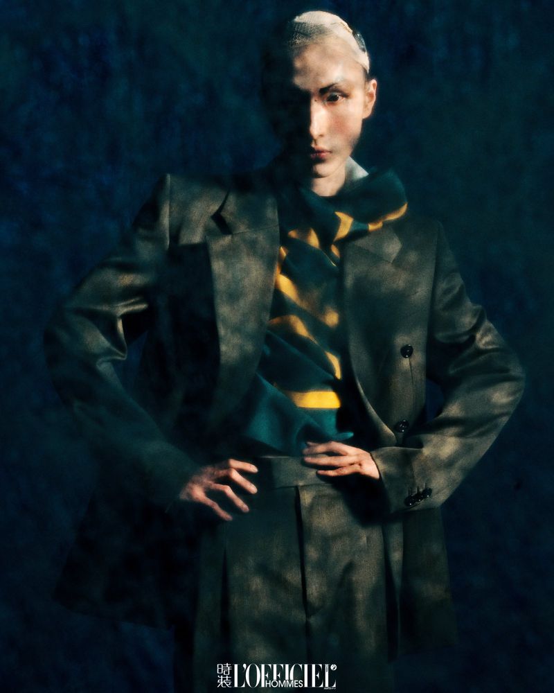 Fashion editorial portrait featuring a person with a shaved head wearing a dark suit and a striped scarf against a dark blue background.