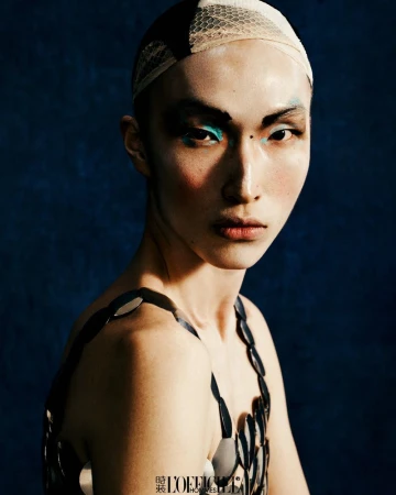 A portrait of an adult with blue makeup on the face, wearing a head covering and disc-shaped top, posing against a blue background.