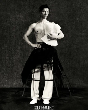 Shirtless male model wearing a sheer black veil, crinoline cage skirt, and white trousers against a textured dark backdrop