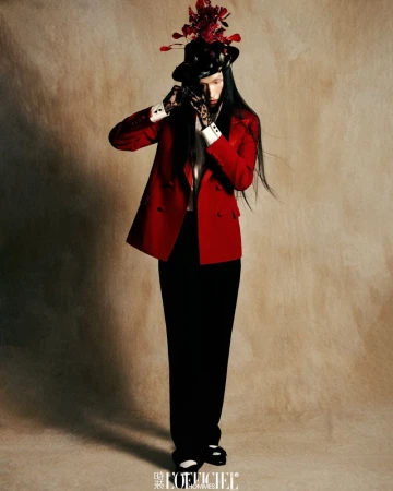 Model in red blazer and black trousers wearing a floral hat and lace gloves against a mottled brown background