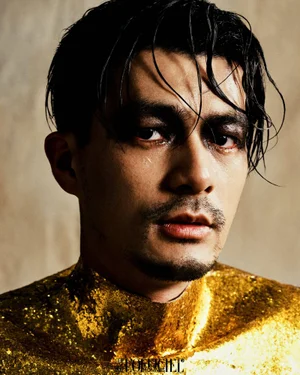 Close-up portrait of a man with wet dark hair and dramatic dark eye makeup wearing a glittering gold top against a neutral textured wall.