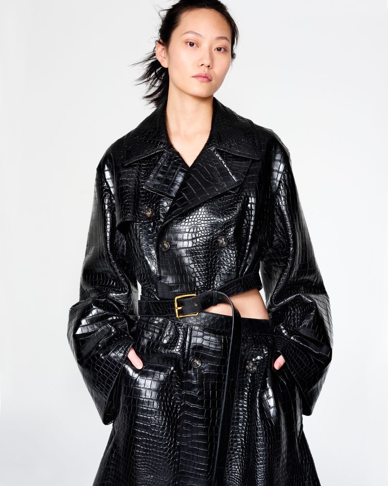 An Asian woman models a double-breasted, black, crocodile-texture trench coat with a belt and matching skirt against a white backdrop.